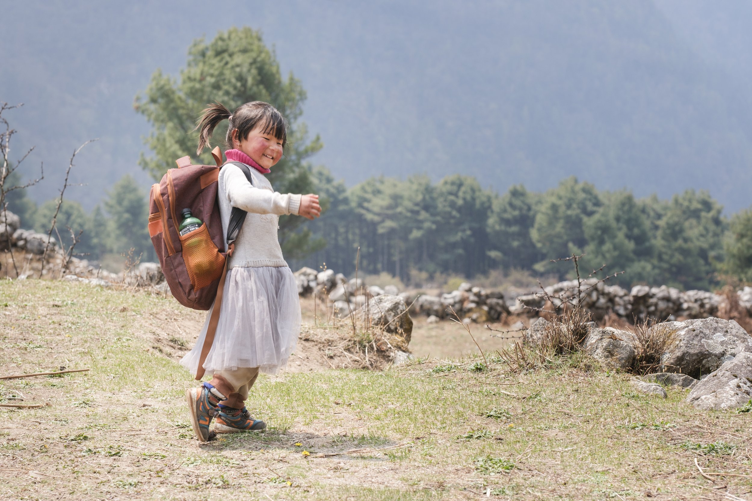 A young girl with dark hair tied in a ponytail, wearing a pink sweater, gray cardigan, and long skirt, walking outdoors with a large brown backpack that has a water bottle in its side pocket, in a landscape with trees and rocks.