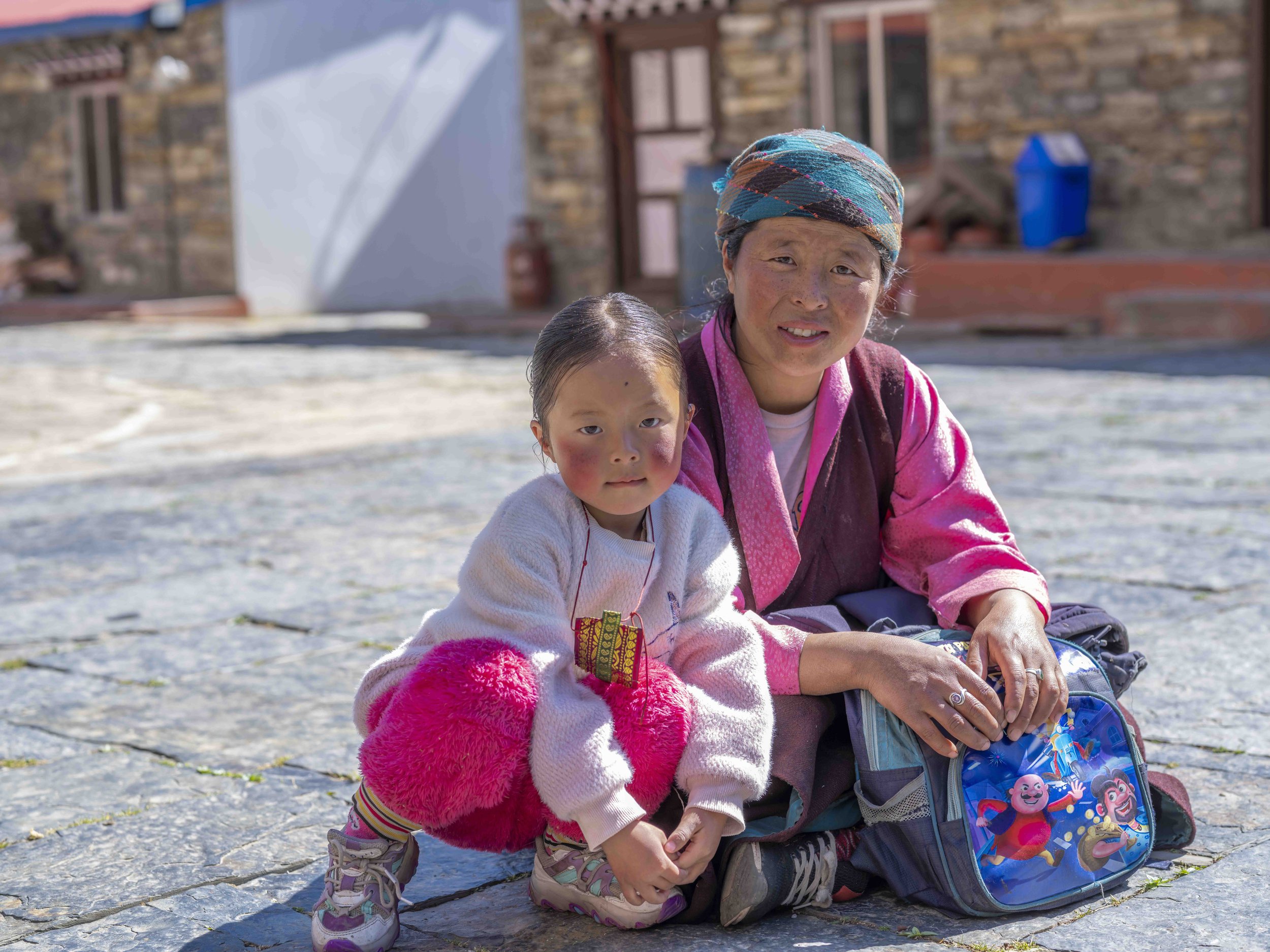 A woman and a young girl sit on the ground outside near a stone building. The woman wears a colorful headscarf and pink clothing, while the girl wears a white sweater and fluffy pink accessories. They are looking at the camera, with a backpack featur