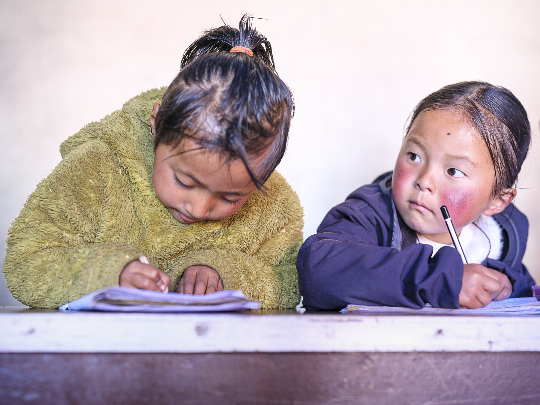 Two young children, a girl and a boy, sitting at a table. The girl is wearing a green fuzzy sweater, looking down at something on the table, with wet hair pulled back in a ponytail. The boy is wearing a dark jacket over a white shirt, holding a pen, and looking to his left, with wet hair and slightly flushed cheeks.