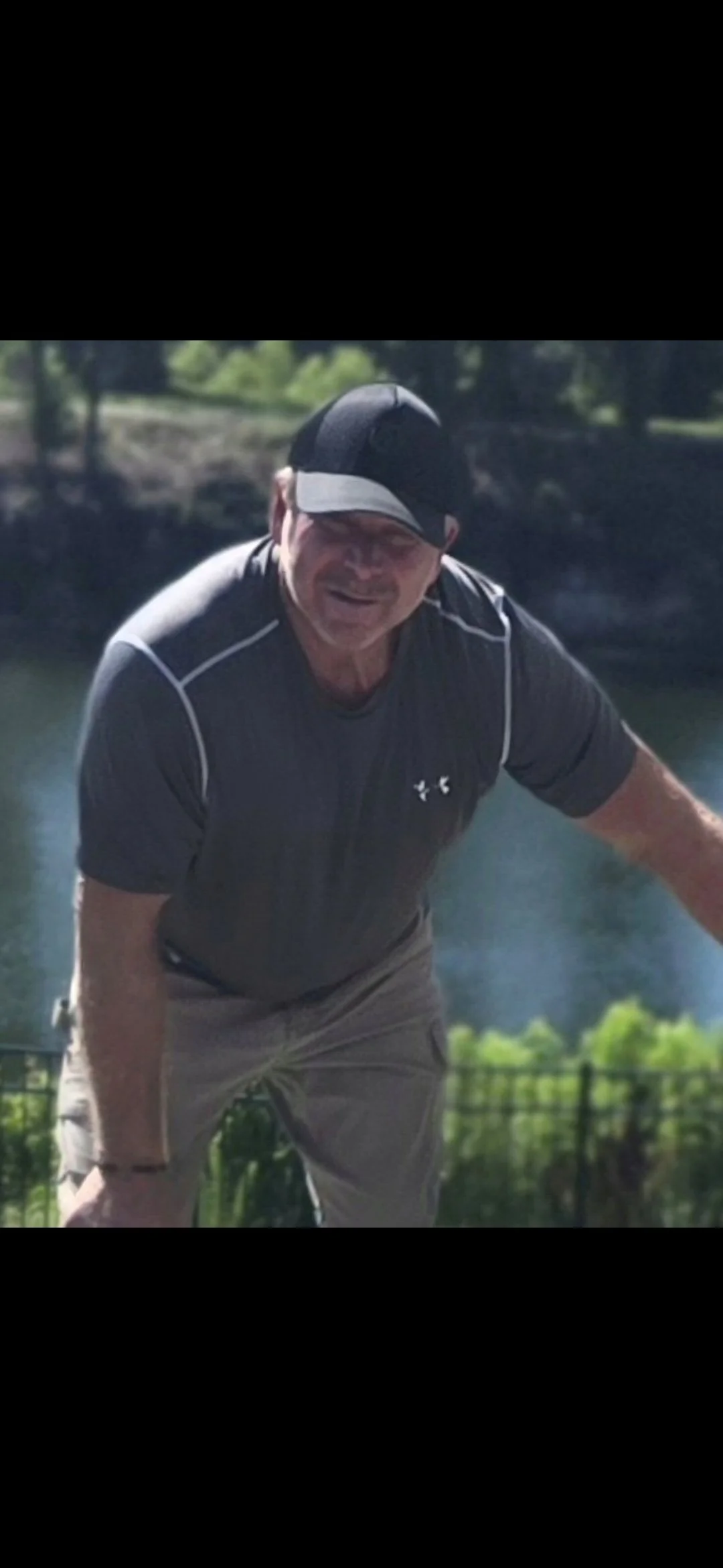 Man smiling outdoors near water, wearing a black cap and athletic clothing.