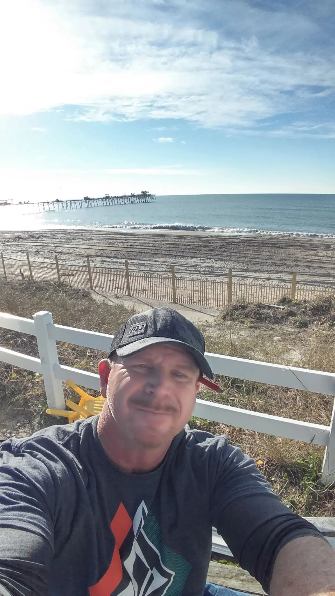 A man taking a selfie at the beach with the ocean and a pier in the background, wearing a black cap and a dark shirt.