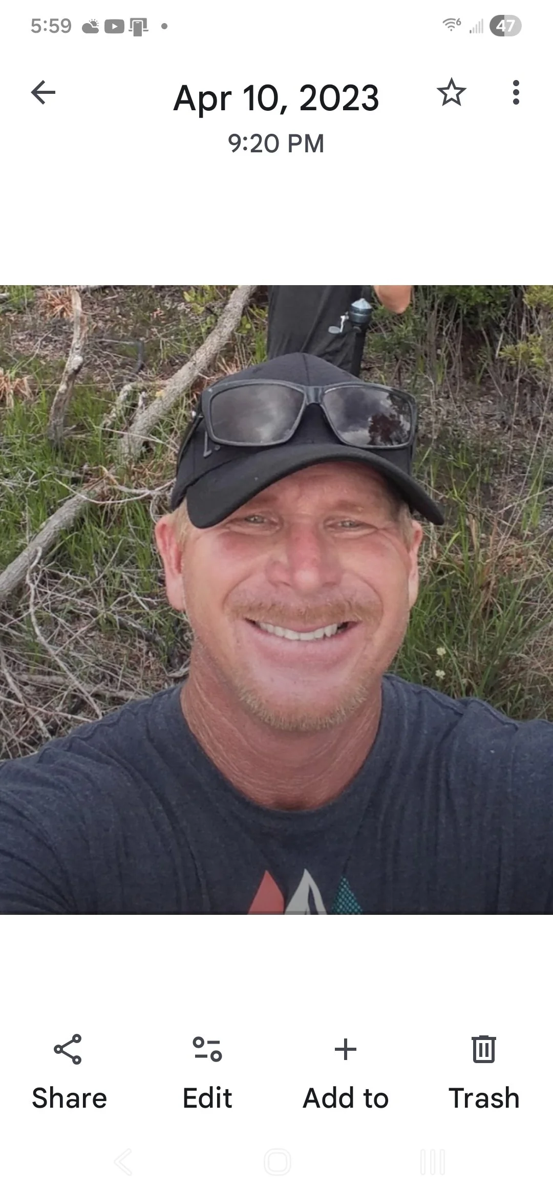 A man smiling in an outdoor setting, wearing a baseball cap and sunglasses on top, with grass and trees in the background.