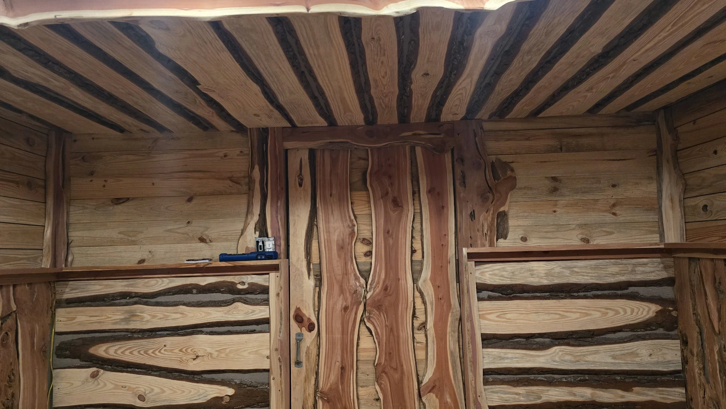 Interior of a wooden room with logs on the walls and ceiling, and a door in the center.