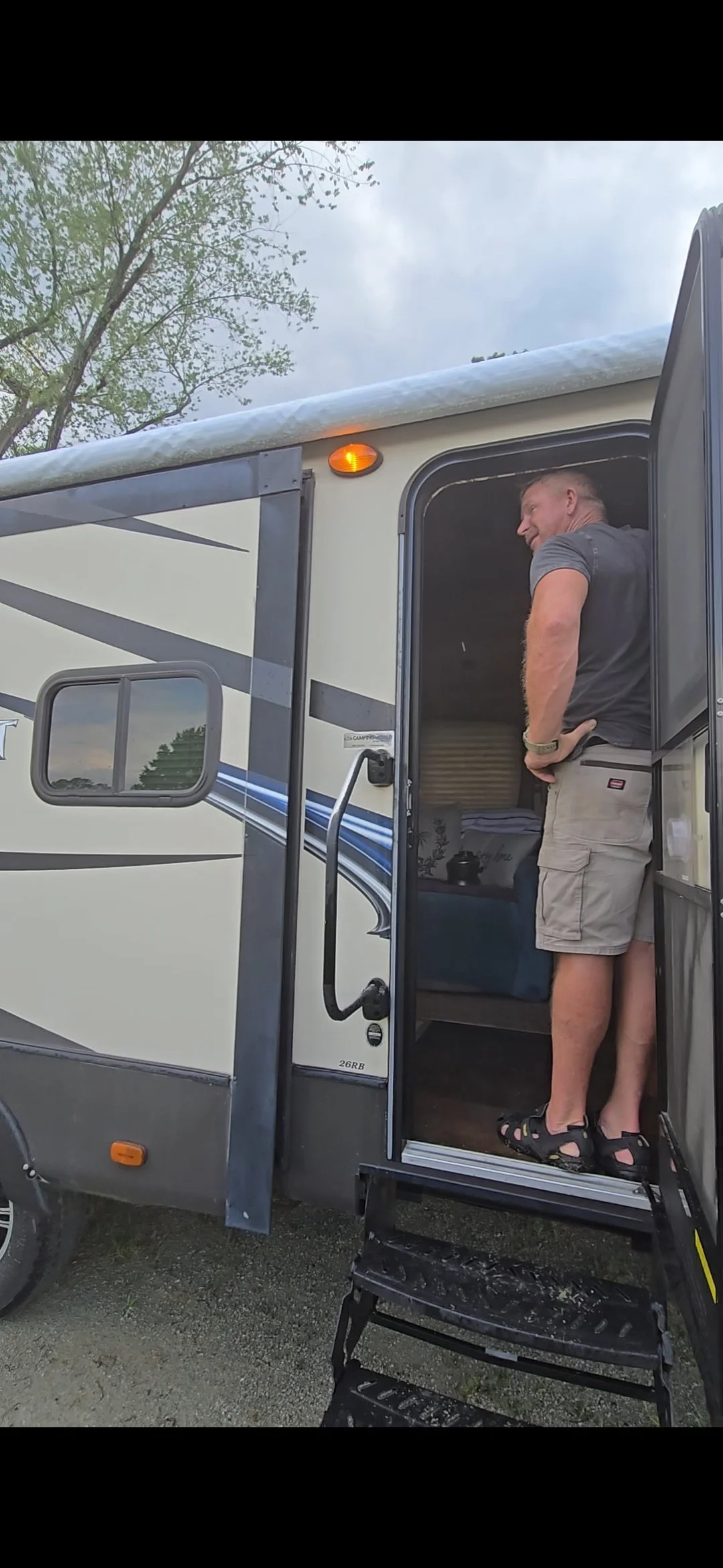 A man standing at the entrance of a travel trailer or RV, looking outside, wearing a gray t-shirt, beige shorts, and sandals, with a tree and cloudy sky in the background.