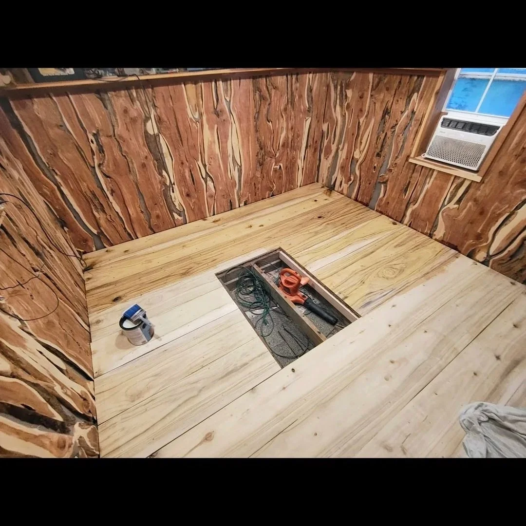 Room with wooden walls and floor, a central cutout in the floor with tools inside, a tape measure and a power saw nearby, and an air conditioning unit in the window.