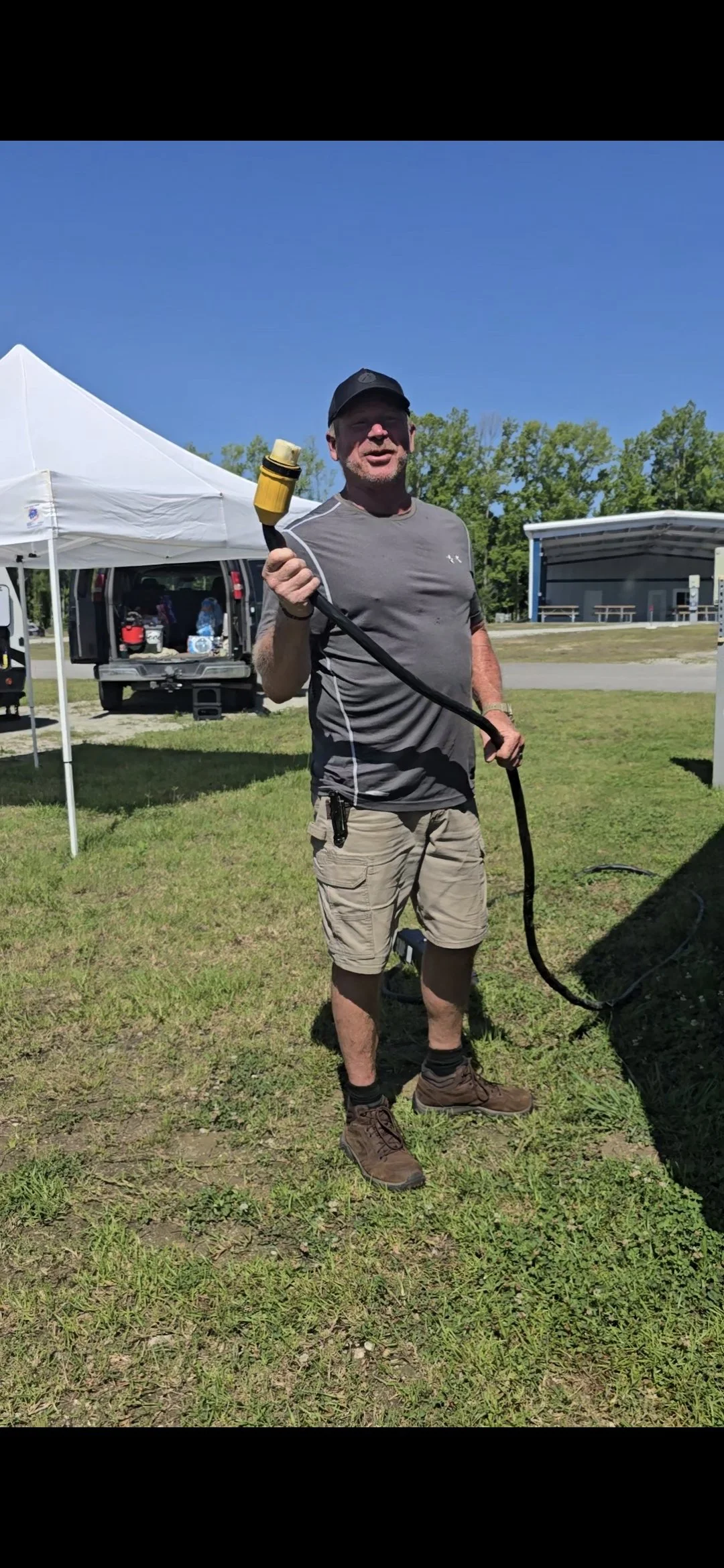 A man holding a yellow garden hose outdoors on a sunny day, with tents and a stage in the background.