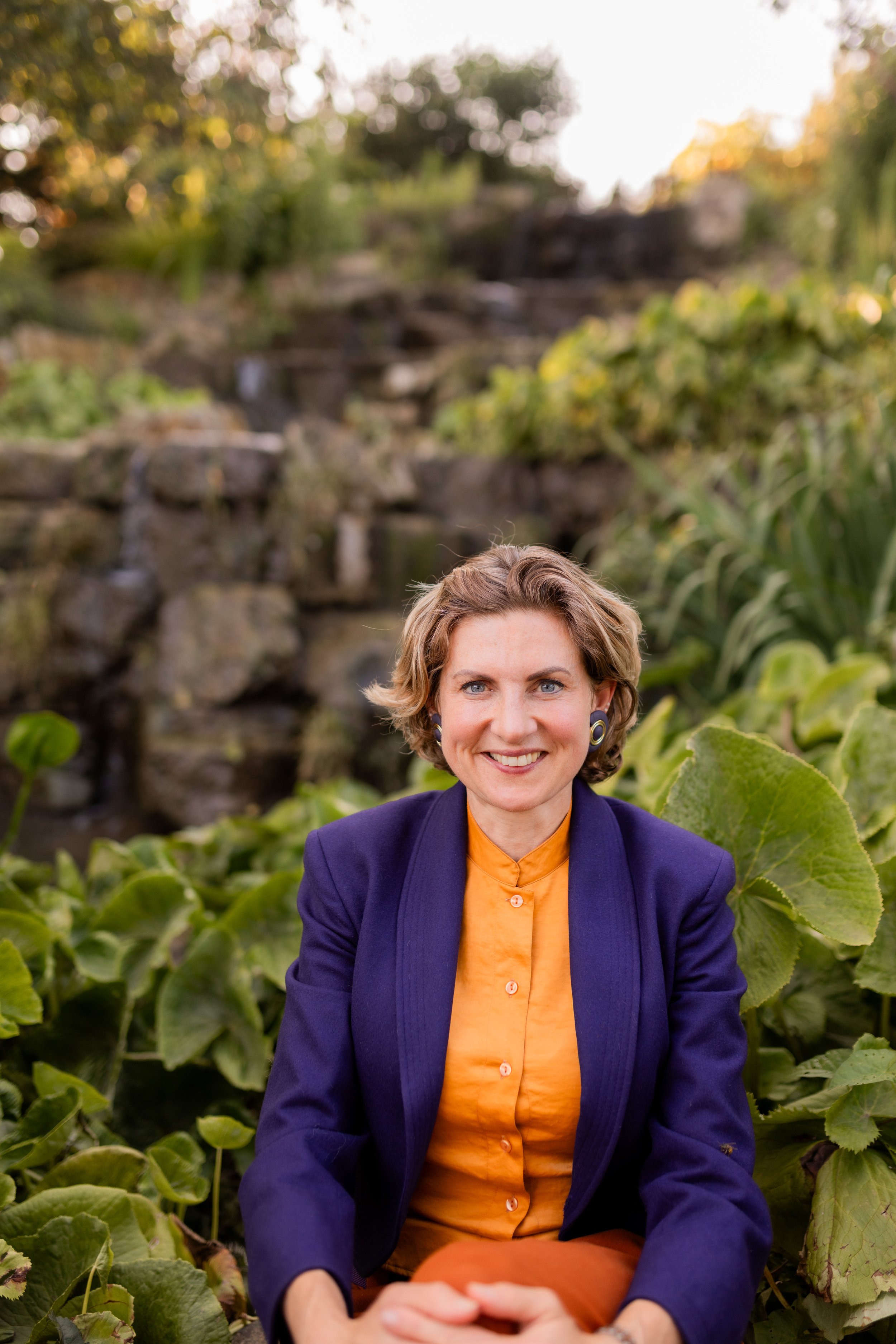 A woman with short brown hair, blue eyes, wearing a purple jacket and an orange shirt, sitting outdoors among green plants with a blurred rocky waterfall background.