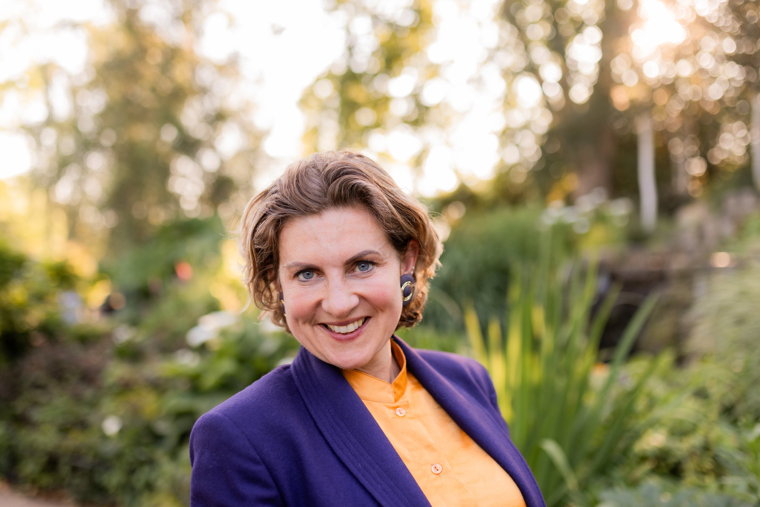 Headshot of a woman with short curly hair smiling outdoors, wearing a purple blazer and yellow top, with a background of trees and sunlight.