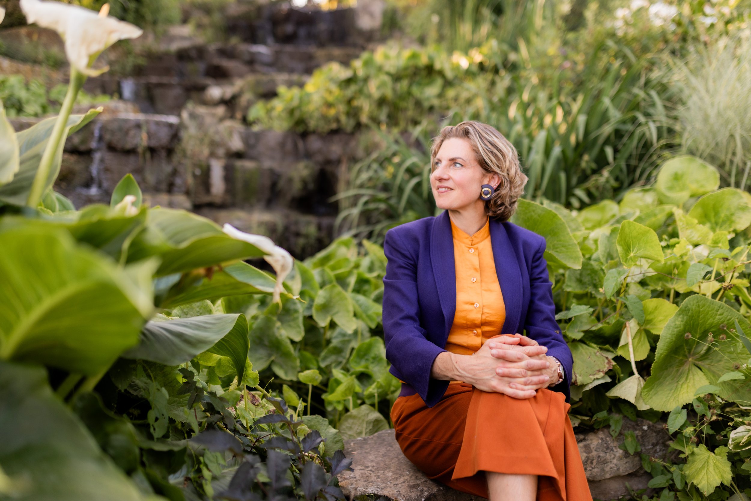 A woman with blonde curly hair sitting outdoors on a rock surrounded by green plants and foliage, with a waterfall in the background, wearing a blue blazer, orange blouse, and orange skirt.