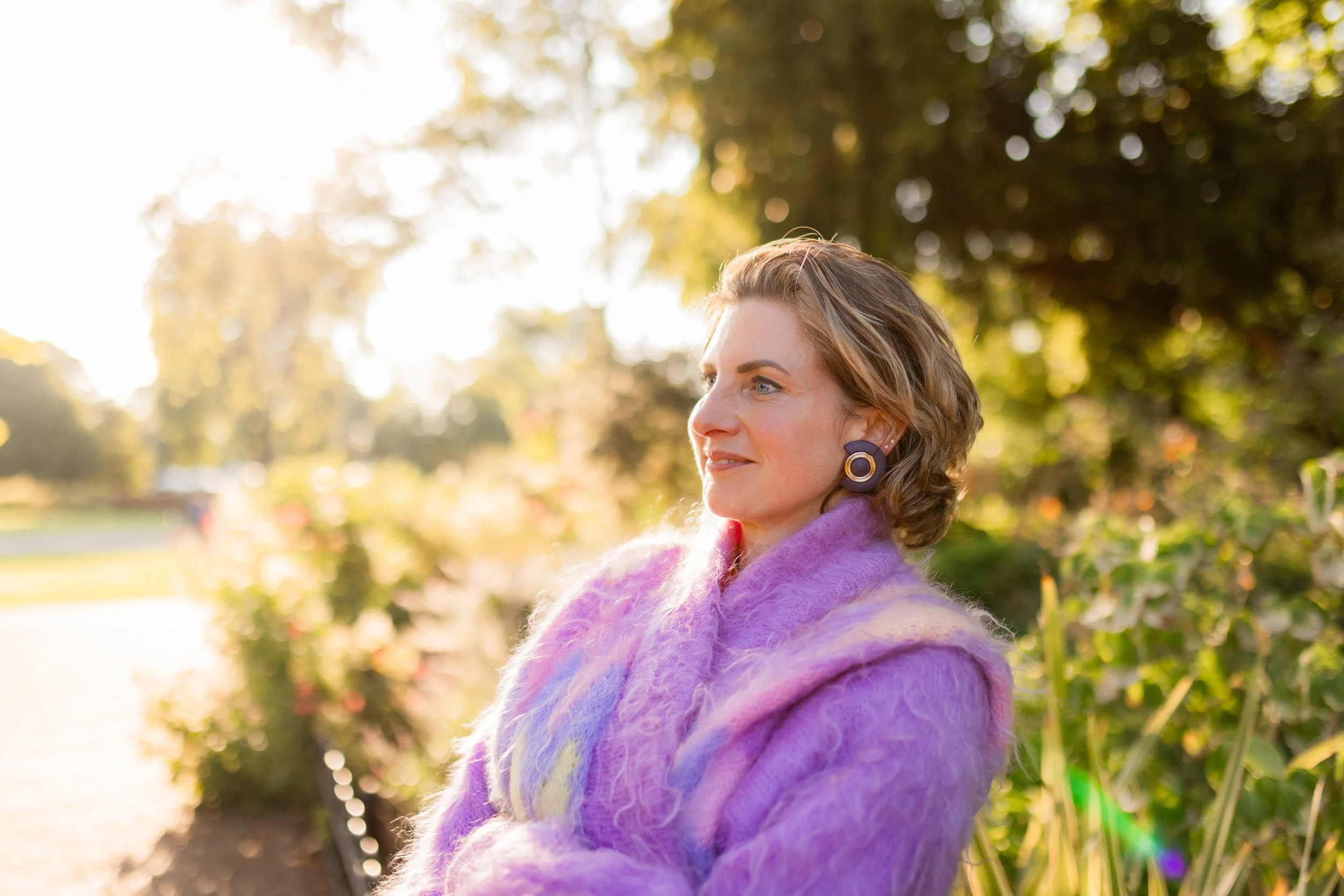 A woman with curly light brown hair wearing a purple fuzzy coat and large hoop earrings standing outdoors in a park with green trees and sunlight in the background.