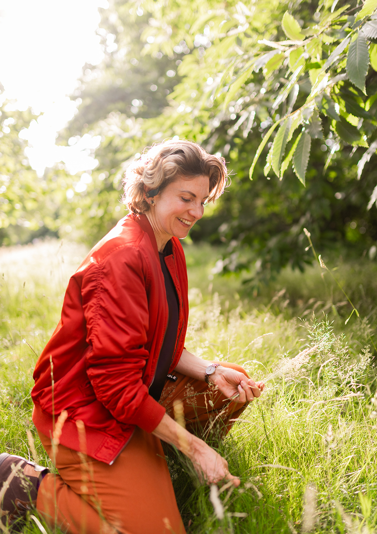 A woman in a red jacket kneeling in a grassy field, smiling while picking plants, with trees and sunlight in the background.