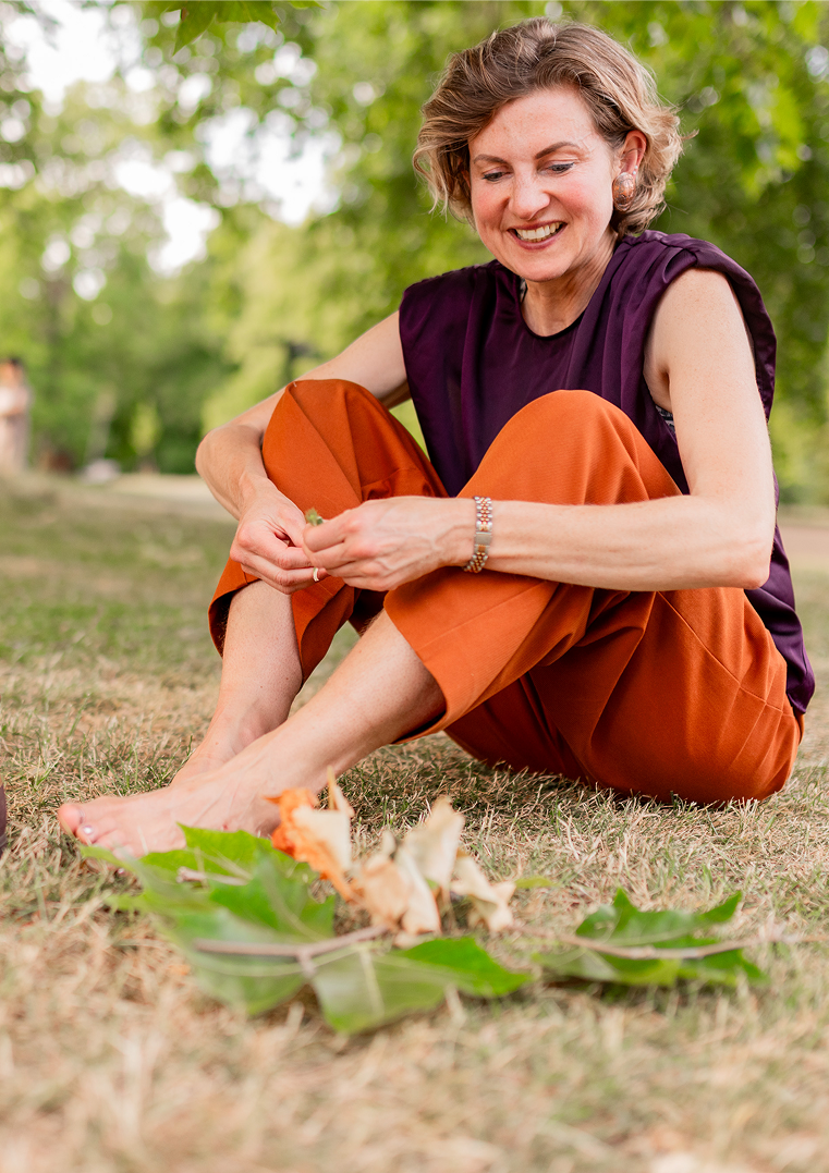 A woman sitting on the grass outdoors, smiling while peeling a carrot. She is wearing an orange skirt and a purple sleeveless top, with trees in the background.