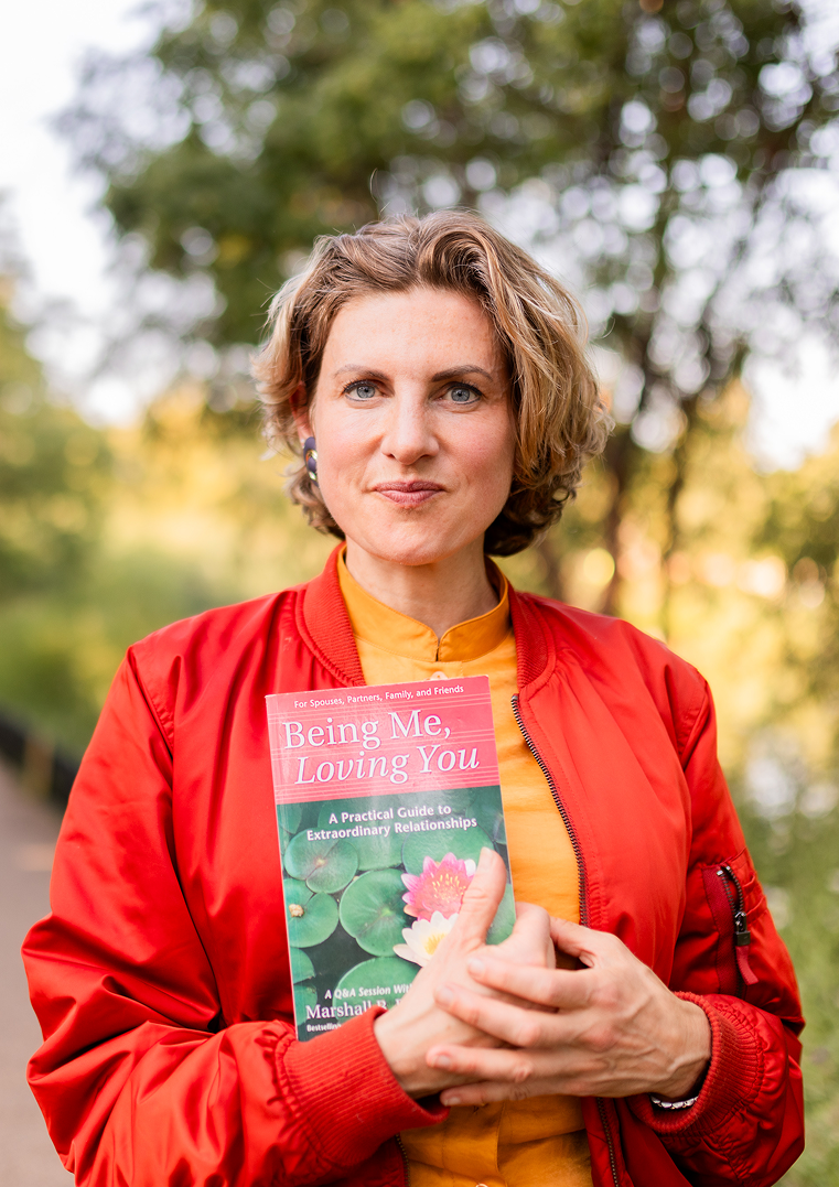 Woman with short curly hair wearing a red jacket and yellow shirt holding a book titled 'Being Me, Loving You' outdoors with green trees in the background.