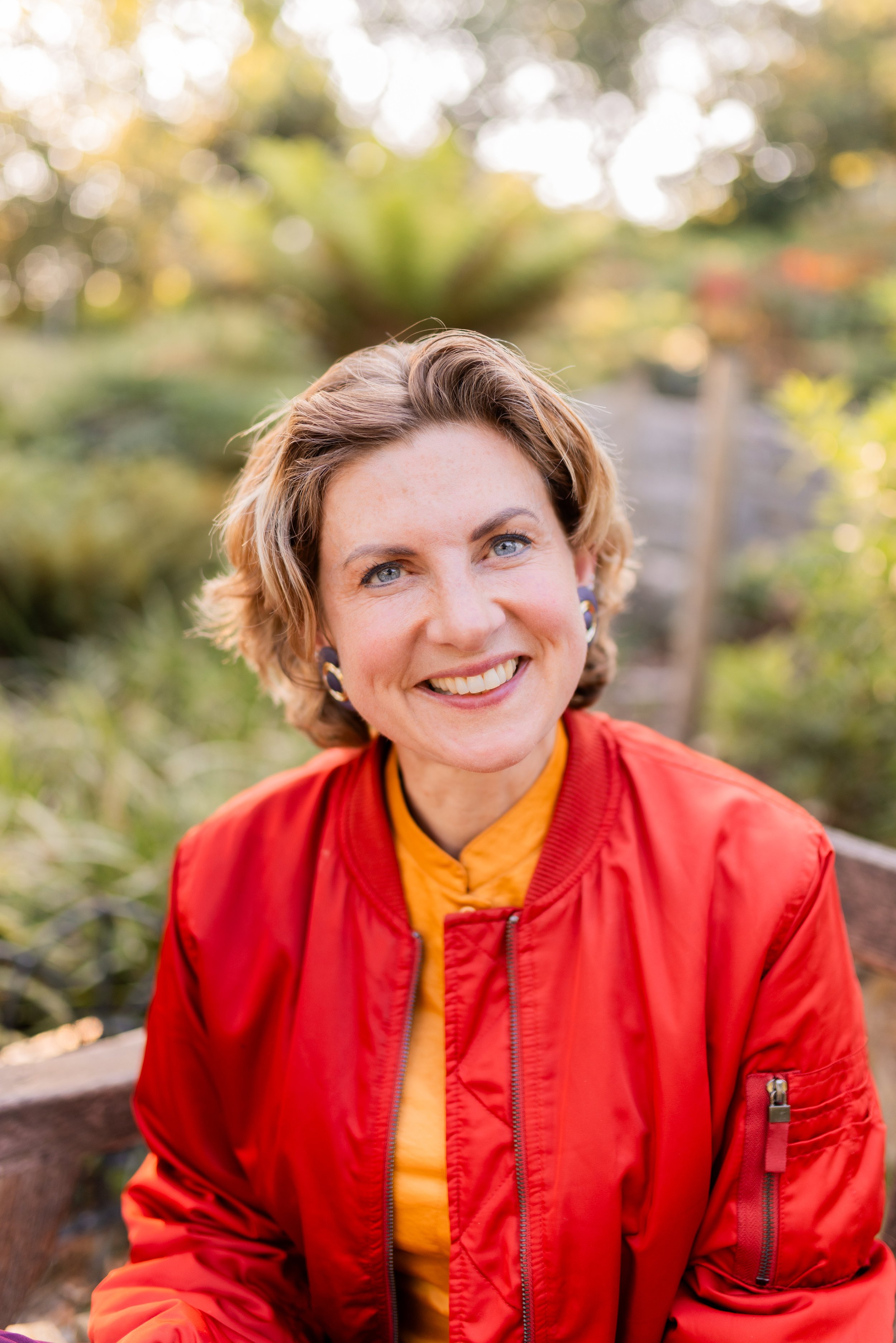 Close-up of a smiling woman with short curly hair wearing a red jacket and yellow shirt, outdoors with greenery and sunlight in the background.