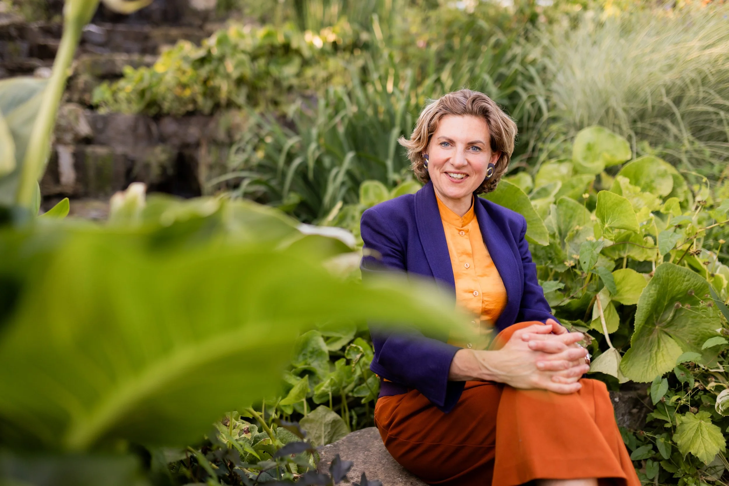 A woman with short blonde hair, wearing a dark blazer and bright orange blouse, sits outdoors among green plants and leaves, smiling at the camera.