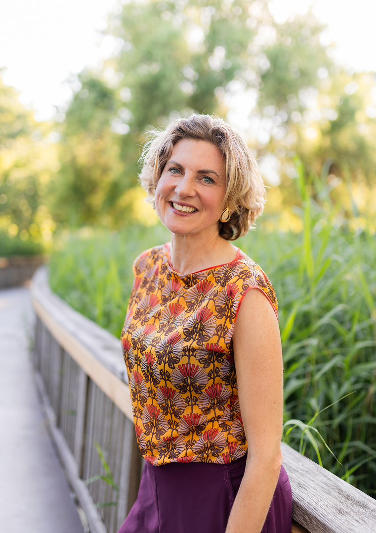 A smiling woman with short wavy blonde hair, wearing a sleeveless floral top in shades of orange, purple, and brown, standing outdoors on a wooden bridge surrounded by greenery.