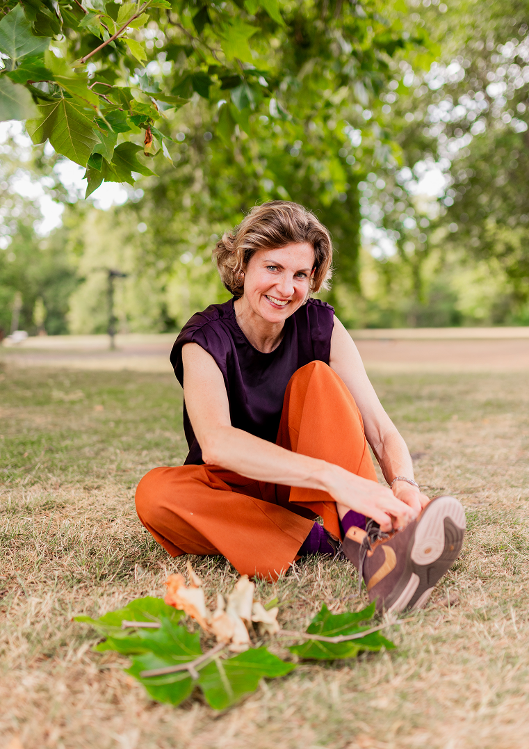 A woman sitting on the grass outdoors under a tree, smiling while tying her shoelace. In front of her, there are some scattered leaves and small branches.