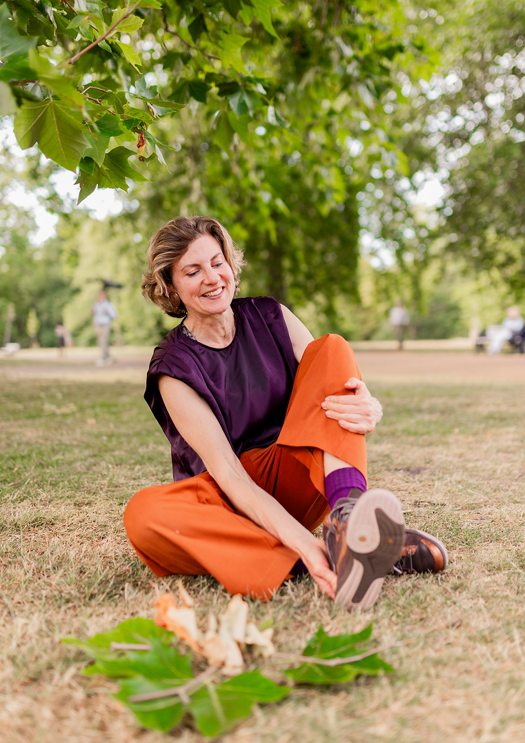 Smiling woman sitting on the grass, adjusting her shoe, with green trees and leaves overhead, in a park.