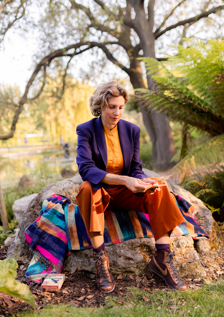 A woman sitting outdoors on a rock, reading a book, with a colorful blanket and a box of Urban Crow on the ground beside her, surrounded by trees and greenery.