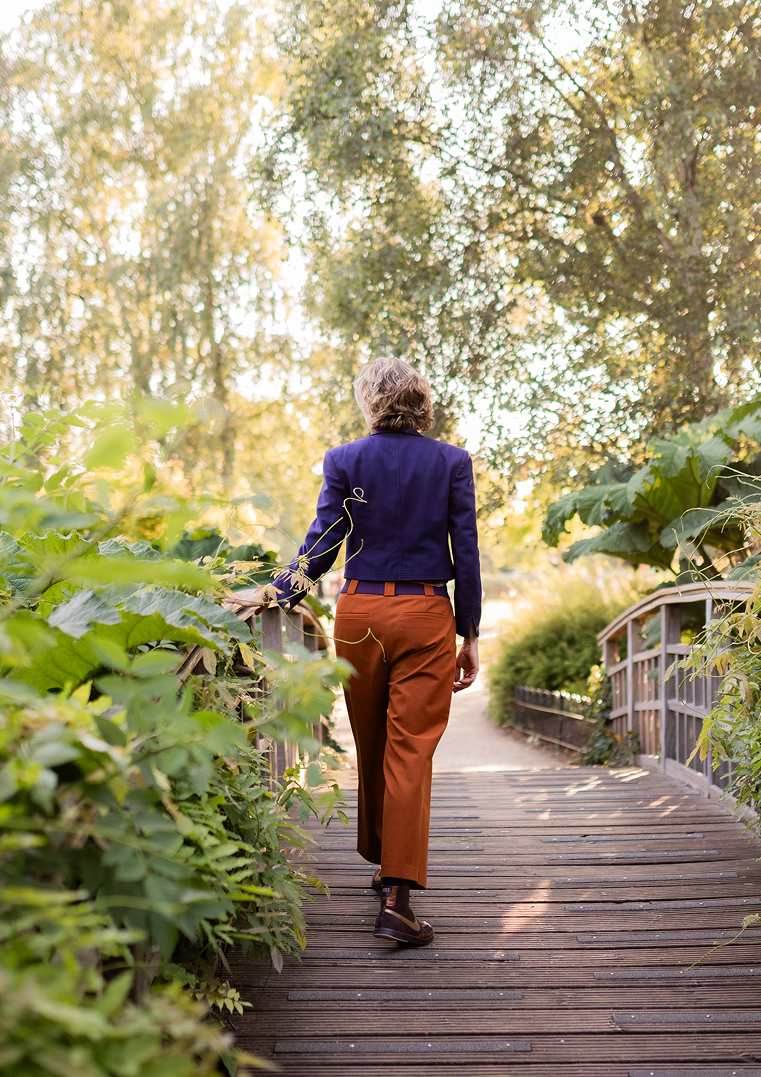 A woman walks on a wooden bridge surrounded by lush green foliage and trees, with sunlight filtering through the leaves.