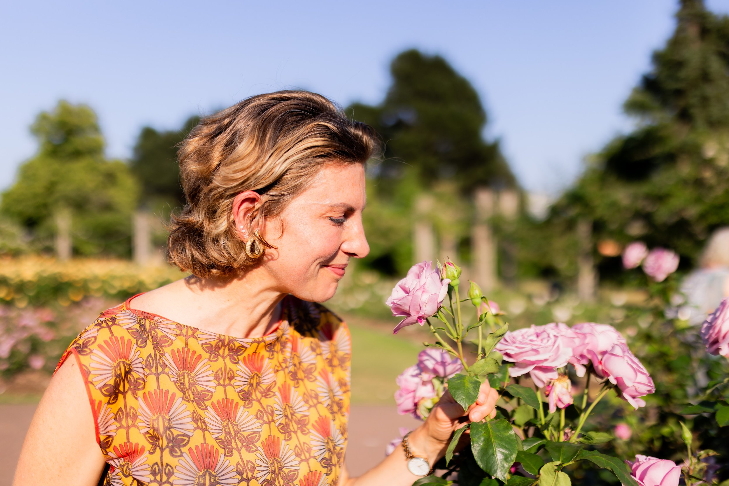 A woman with short brown hair and earrings holds pink roses in a garden, smiling with eyes closed on a sunny day.