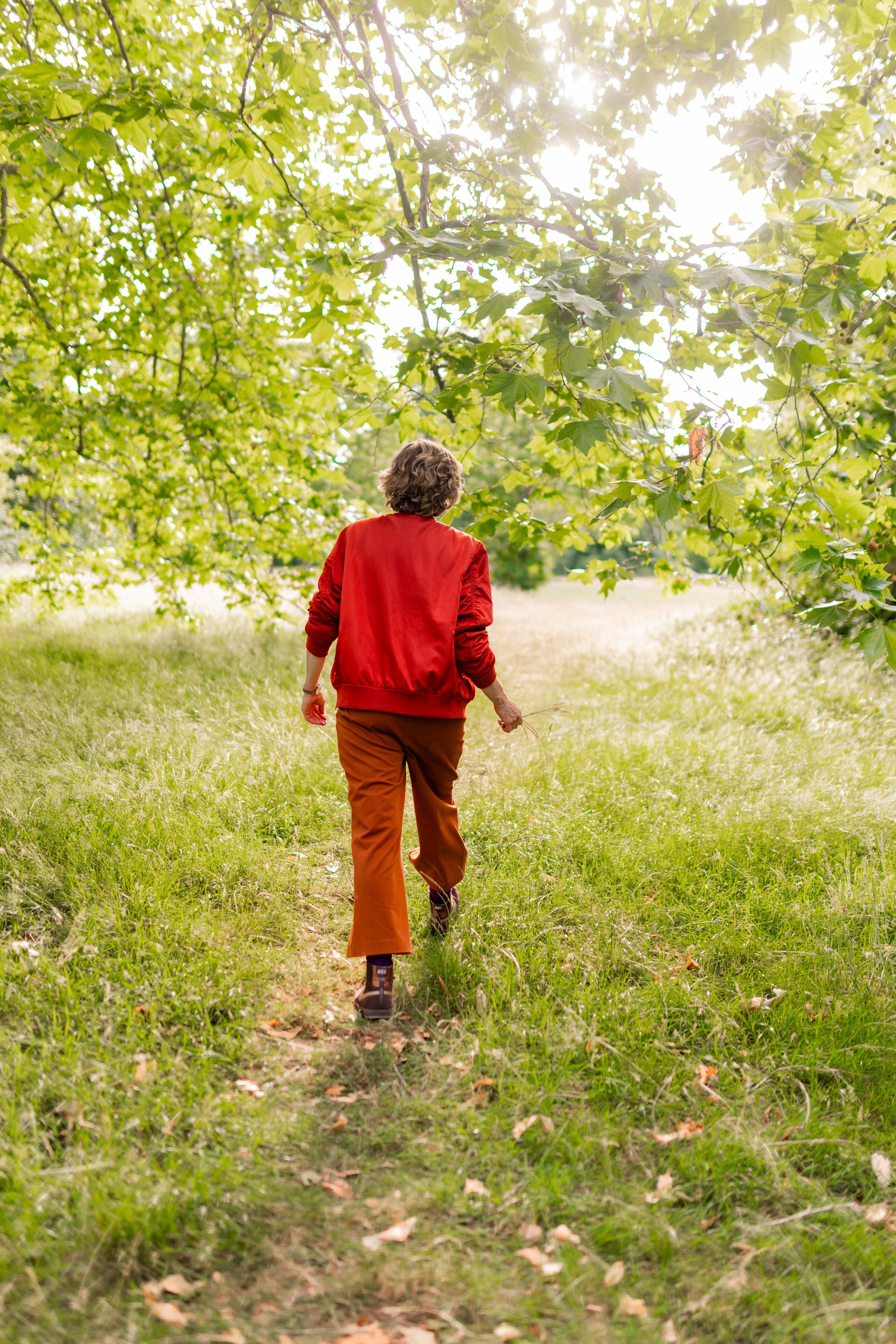 A person with short curly hair wearing a red jacket and brown pants walking on a grassy trail under green leafy trees during daytime.