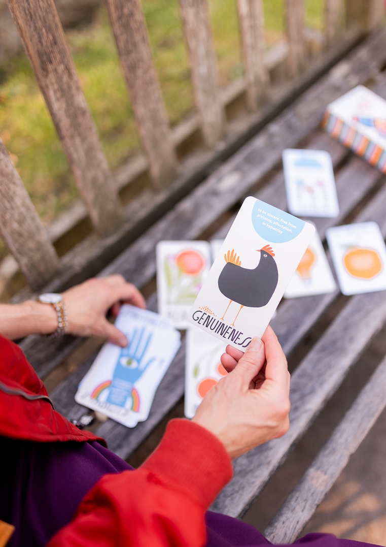 Person holding a card with a black chicken illustration and the word 'Genuineness', with other cards on a wooden bench.