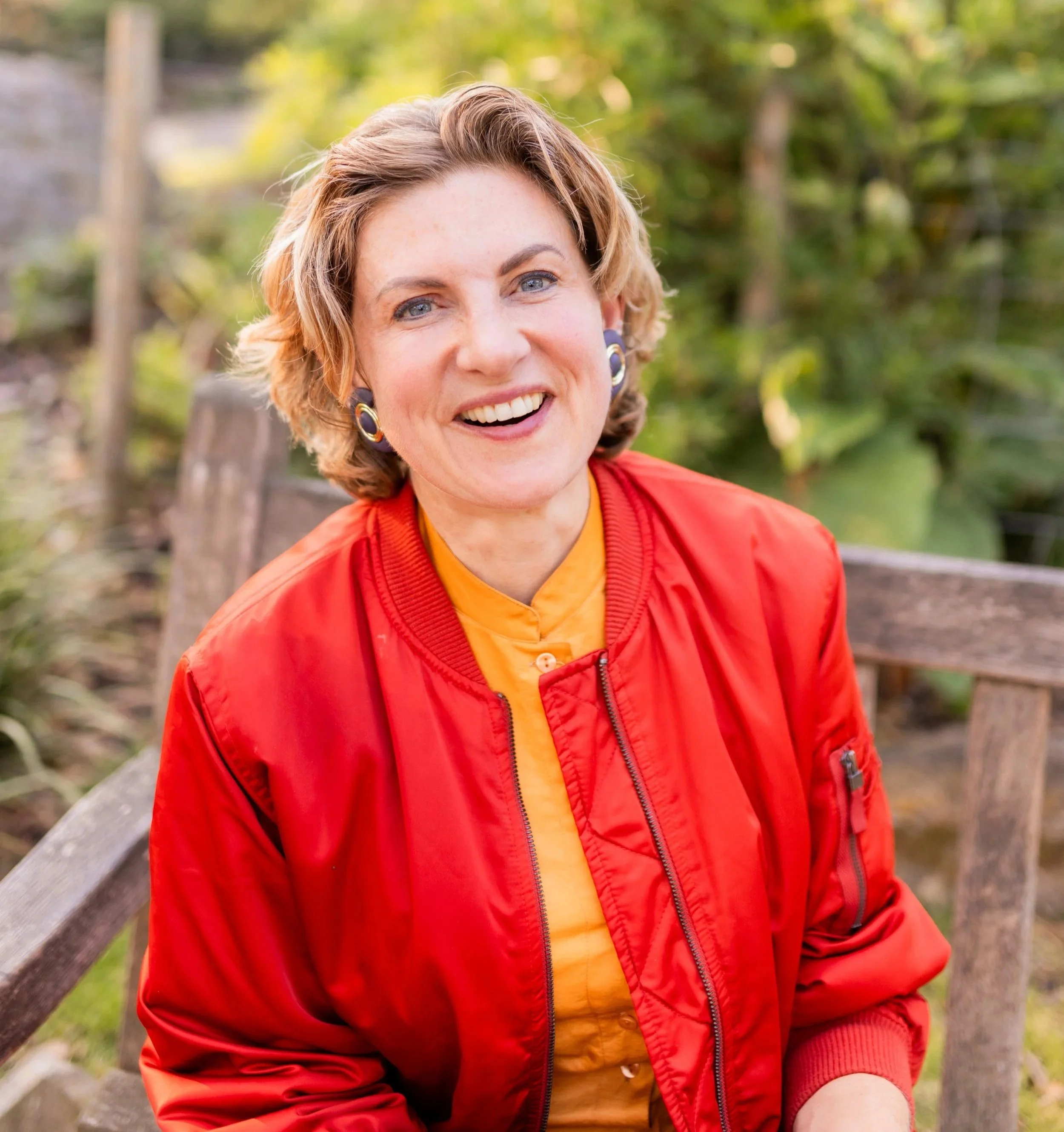 A smiling woman with short, curly blonde hair, blue eyes, wearing a red jacket and yellow shirt, sitting on a park bench surrounded by green trees and foliage.