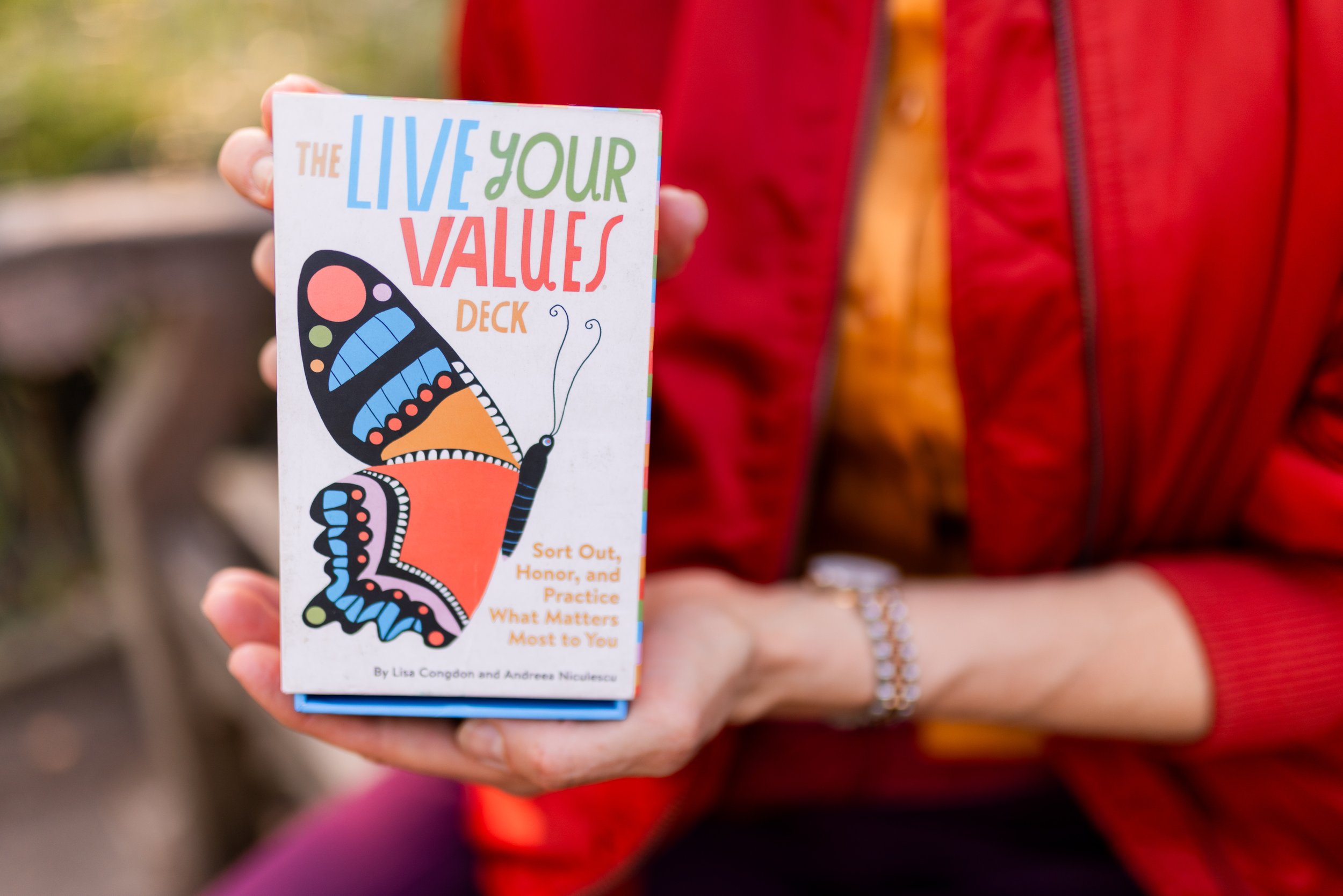 A person in a red jacket holding a colorful booklet titled 'The Live Your Values Deck' with a butterfly illustration on the cover.