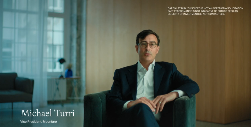 A man in a suit sitting on a green armchair in a modern, well-lit office or living room with a wooden wall and large window in the background.