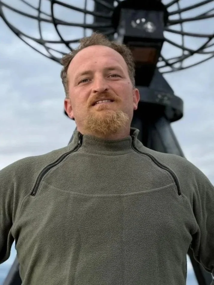 Kuvassa TYYNI Remontit & Huollot Oy:ssä työskentelevä tiiminvetäjä ja rakennustyöntekijä Valmir hymyilee, taustalla on vanha radioastronominen teleskooppi joka kuvaa, että Valmir on myös työssään tarkka.