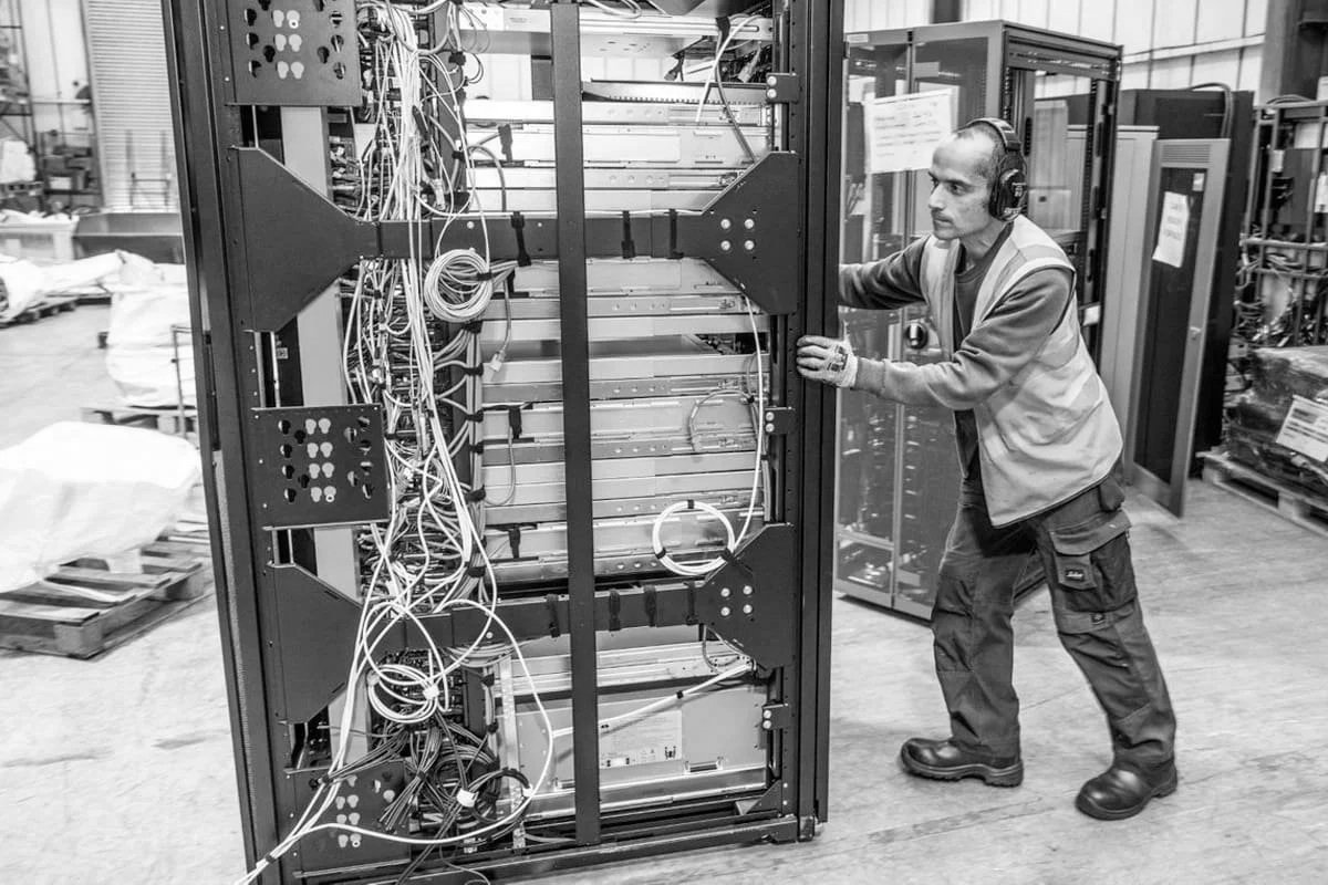 A technician working on a large server rack with open panels, surrounded by cables and electronic equipment in a data center or server room.