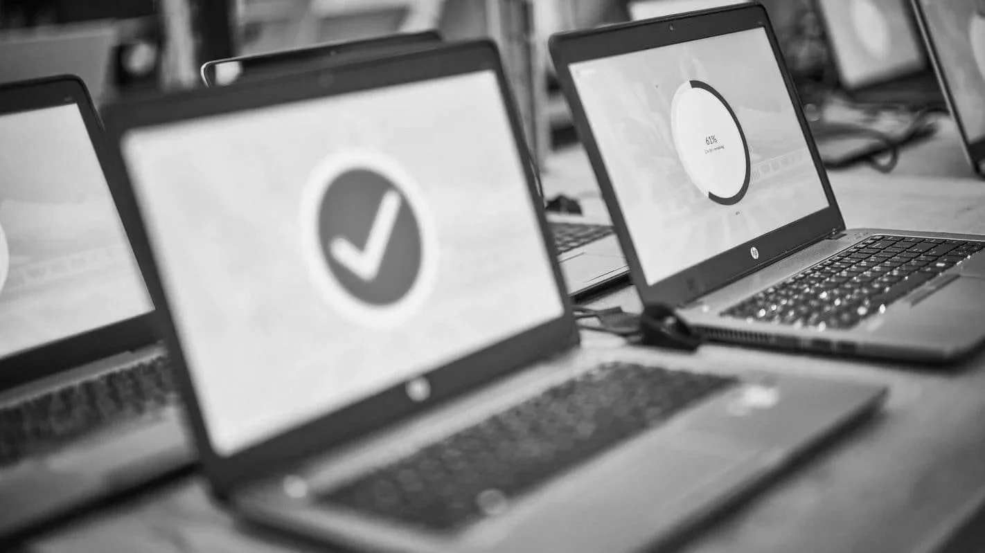Multiple laptops on a table, some screens displaying checkmarks and progress circles, in a grayscale setting.