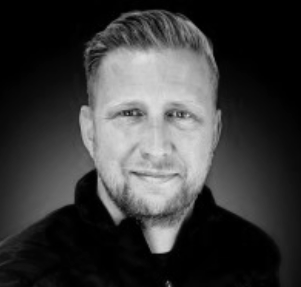 Portrait of a man with short blond hair and a beard, wearing a black jacket, smiling, against a dark gradient background.