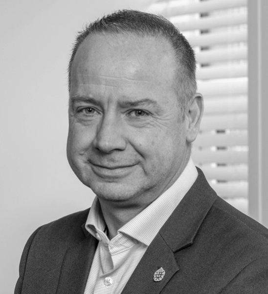 A portrait of a middle-aged man with short light brown hair, wearing a dark suit jacket and a light purple dress shirt, standing in front of window blinds.