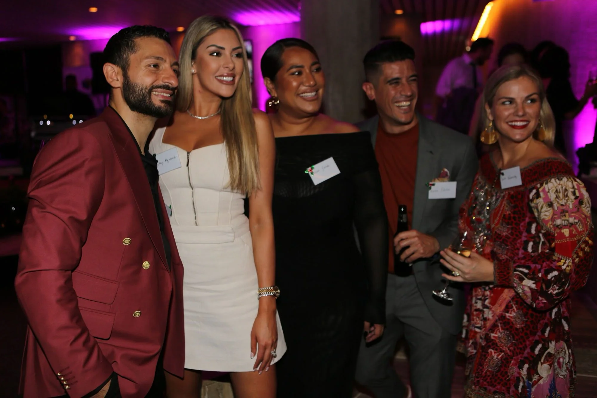 Group of five smiling adults dressed in cocktail attire at a party with pink and purple lighting.