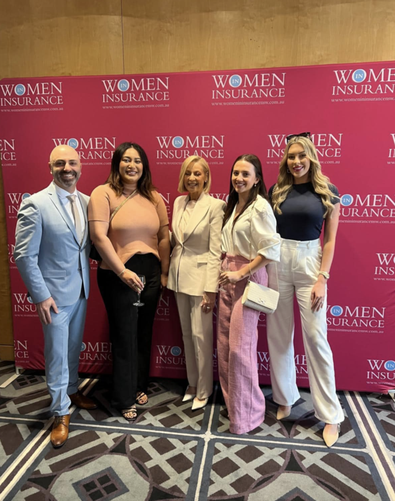 Five people posing in front of a pink backdrop with the words 'Women Insurance' repeated. They are dressed professionally and smiling at the camera.