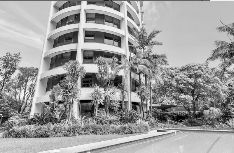 A modern, multi-story building with rounded balconies, surrounded by tropical plants and trees, next to a swimming pool.