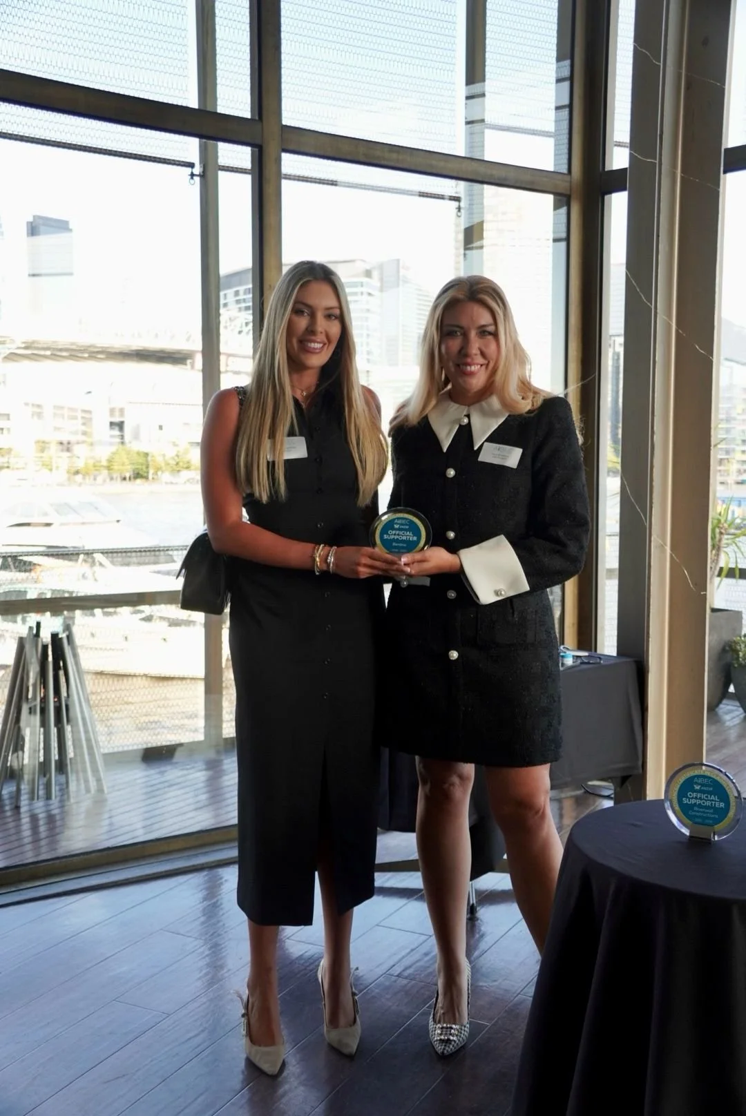 Two women standing indoors holding a circular award plaque with 'Official Supporter' written on it. Large windows with cityscape background. One woman dressed in a black sleeveless jumpsuit, the other in a black blazer dress with white collar and cuffs.