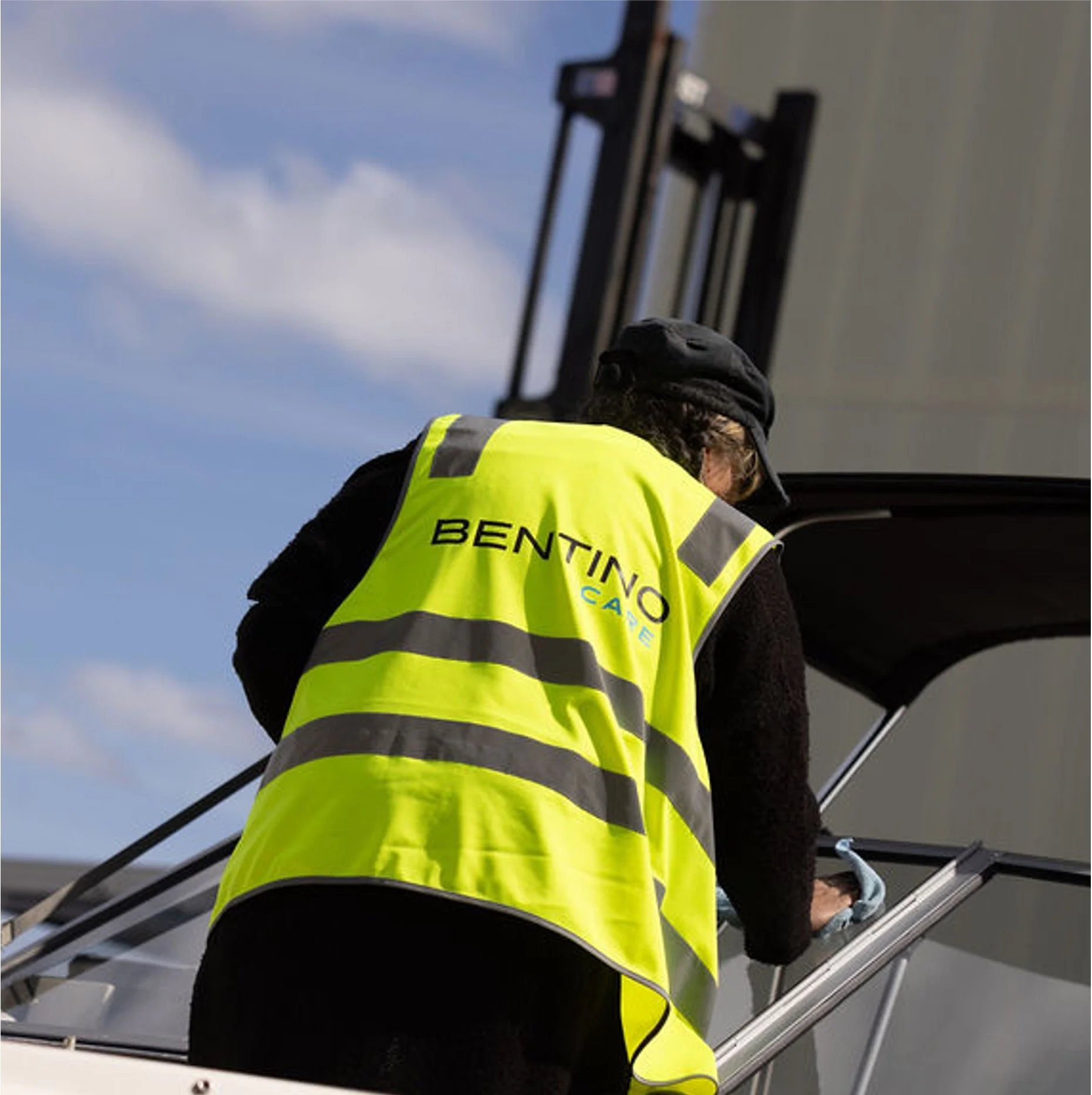A person wearing a high-visibility yellow safety vest with the words 'BENTIGO' visible, working on a boat, using tools or equipment, with a clear blue sky in the background.