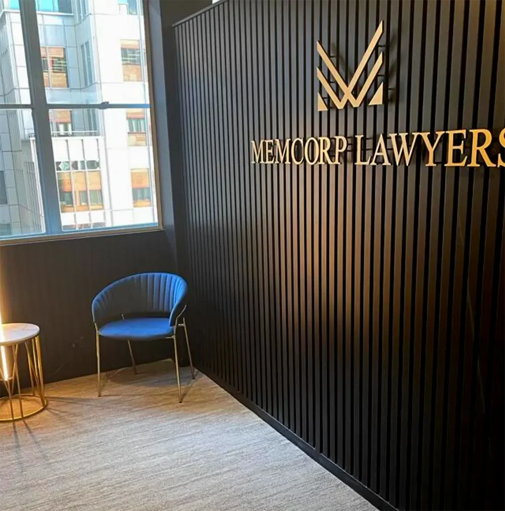 Interior of a modern law office with black textured wall, gold signage reading 'MEMORP LAWYERS', a black velvet chair, and a small side table near a large window showing high-rise buildings outside.