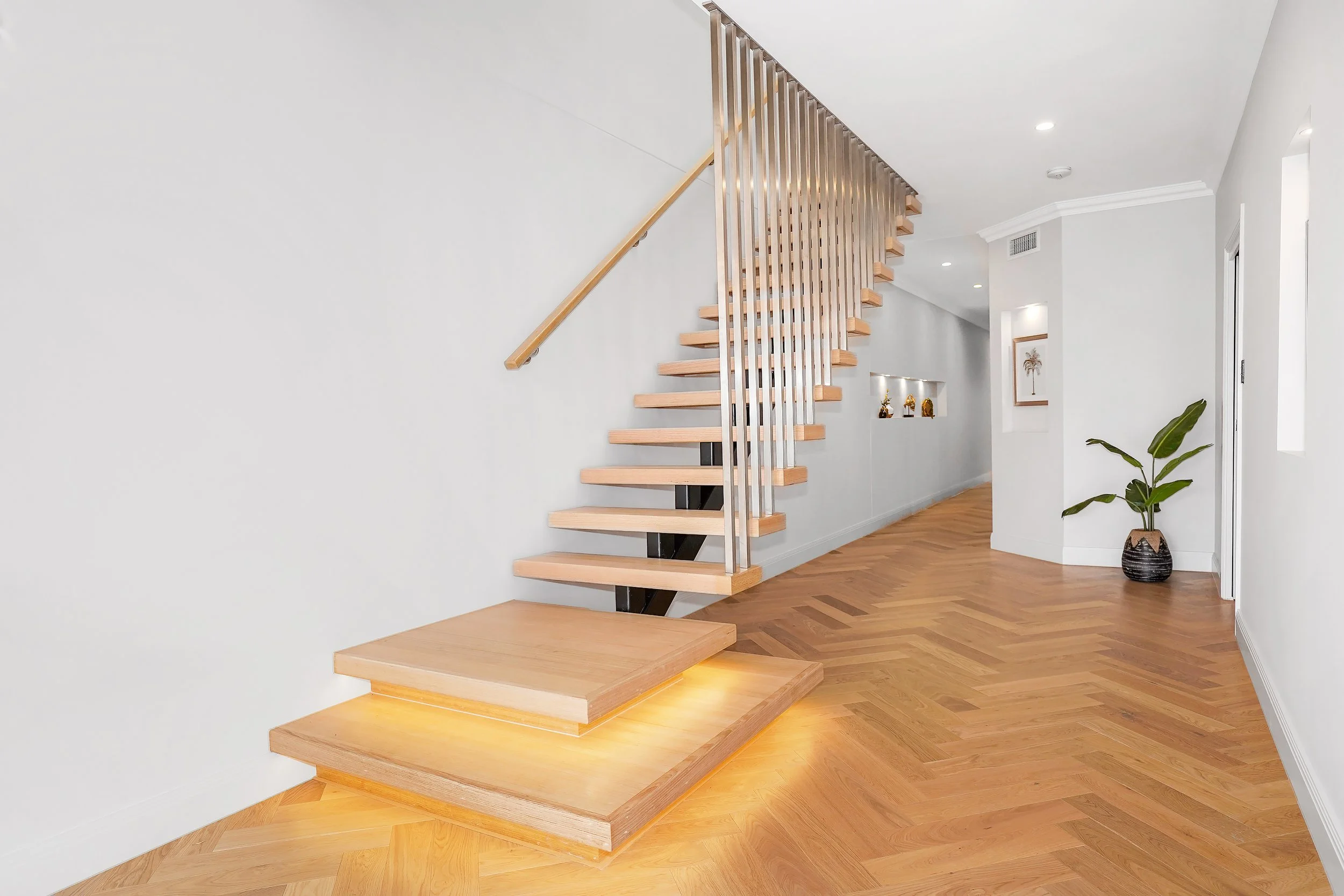 Modern staircase with wooden steps and metal railings in a minimalistic interior with white walls, hardwood flooring, a potted plant, and decorative items on a shelf.