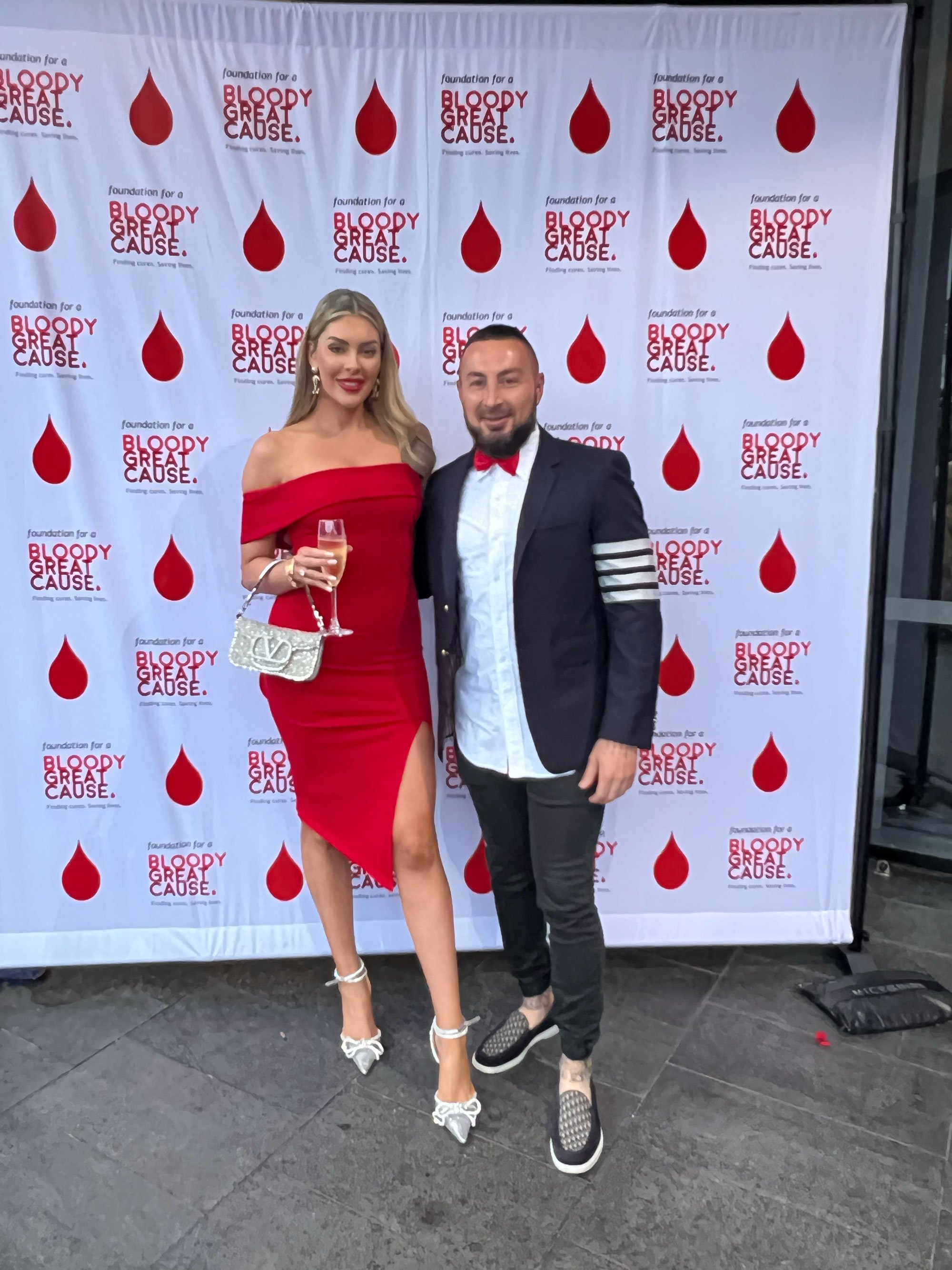 A woman in a red dress and high heels and a man in a suit with a red bow tie stand together holding glasses of champagne in front of a backdrop with blood drop graphics and the text 'foundation for a BLOODY GREAT CAUSE.'