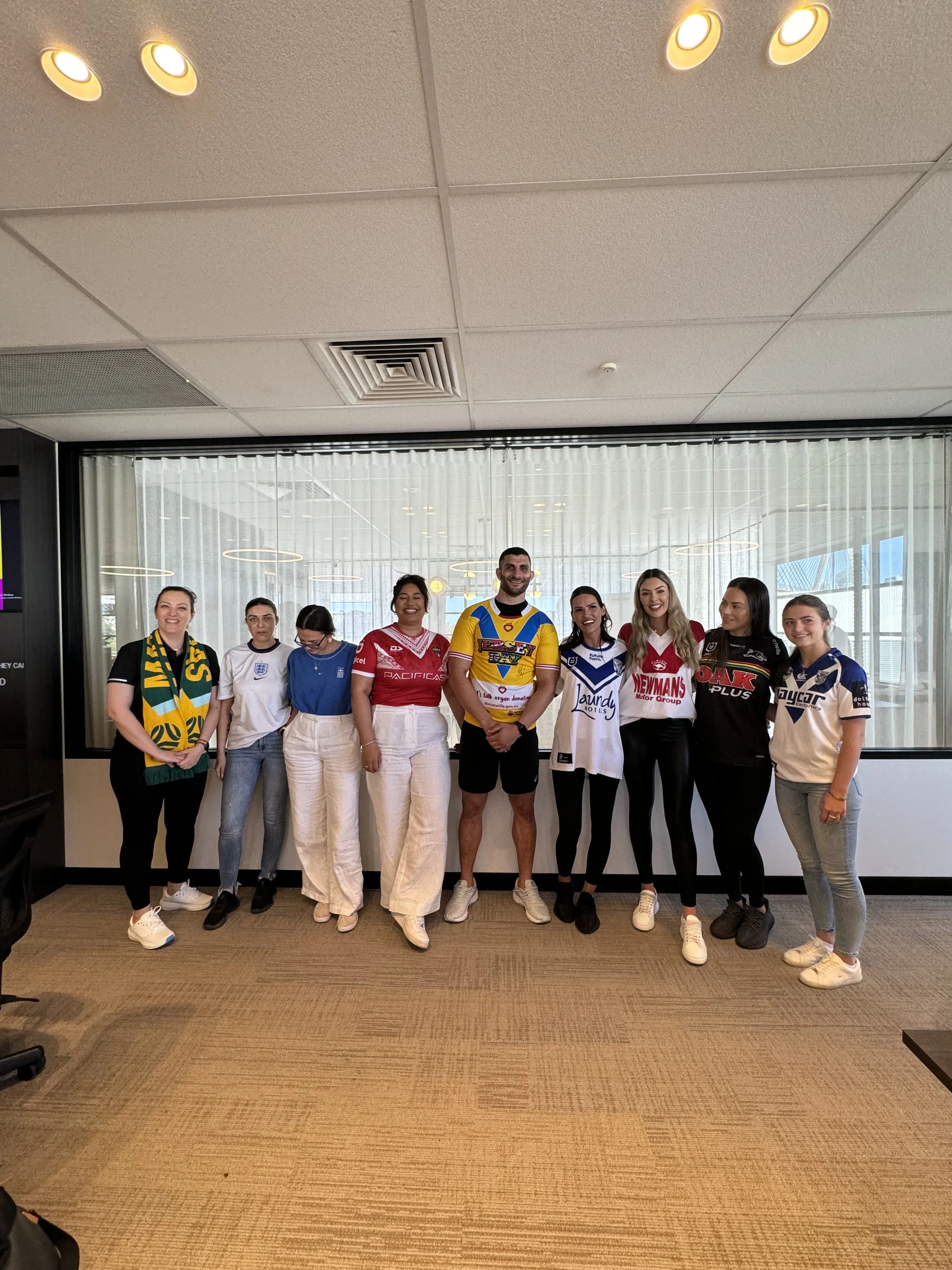 A group of ten people, mostly women, standing in a line indoors in front of large windows with blinds. Some are wearing sports jerseys, and one woman is wearing a green and yellow scarf. The background includes an office-like environment with ceiling lights and a carpeted floor.
