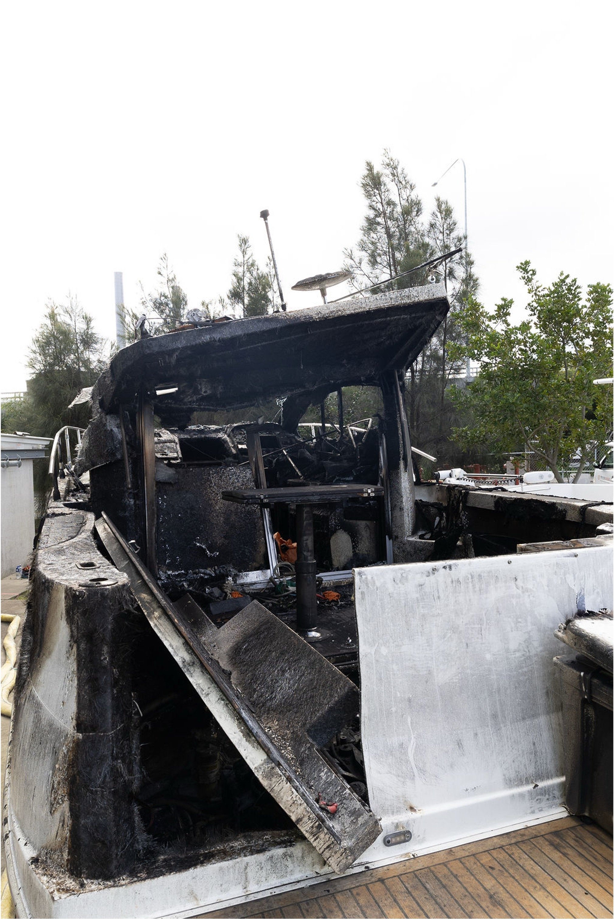 Burned and charred remains of a boat, with black soot and damaged interior, on a wooden dock with trees and a cloudy sky in the background.