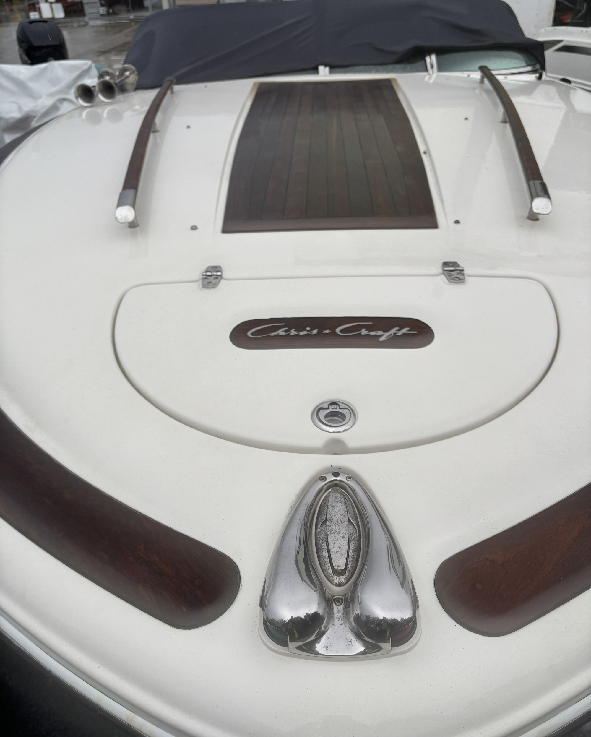 Close-up view of a boat's bow with wood and metal decorations, chrome fixtures, and a black cover in the background.