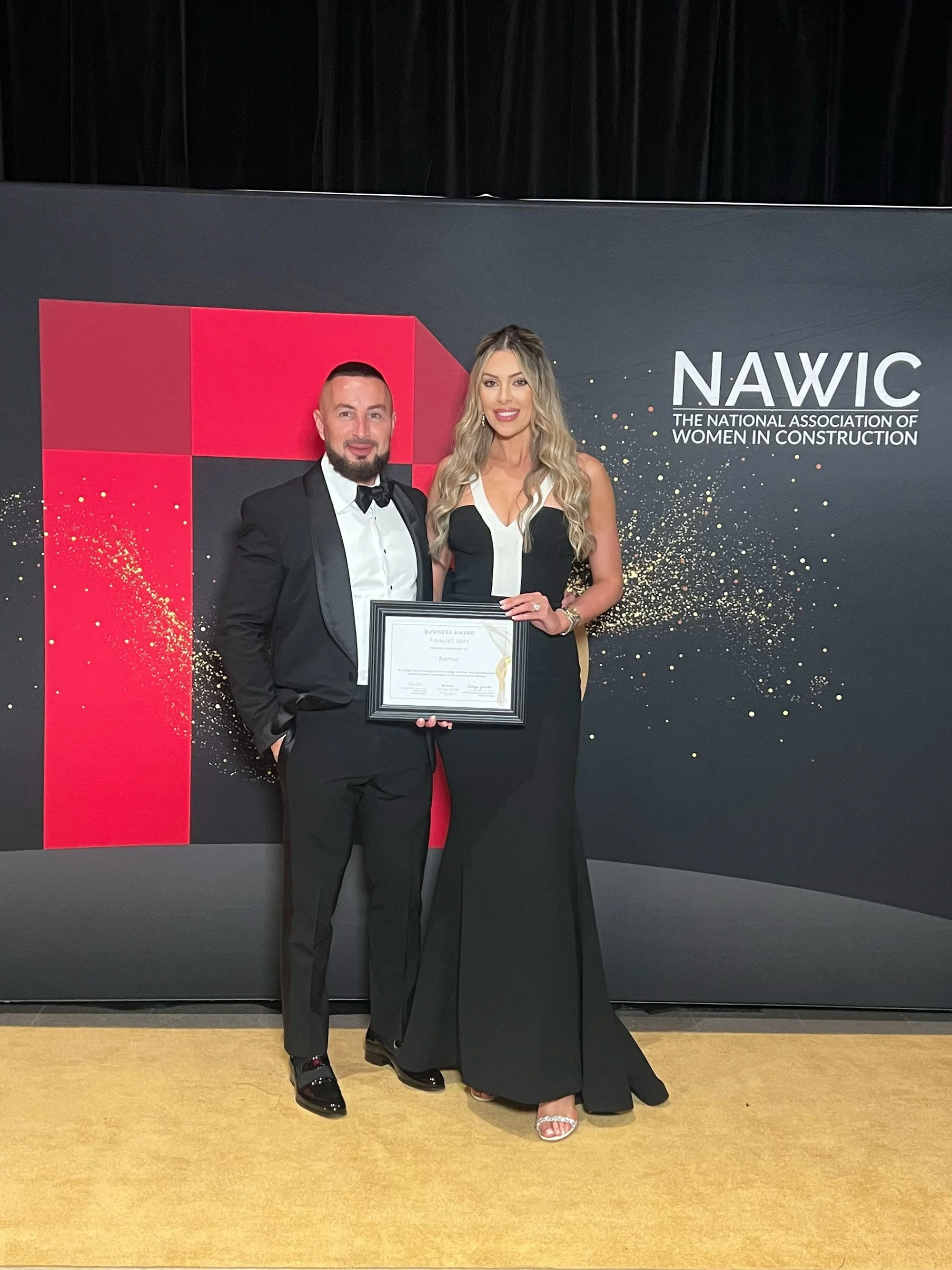 Man in tuxedo and woman in black evening gown holding a framed award at a formal event, standing in front of a backdrop with 'NAWIC' and 'The National Association of Women in Construction' written on it.