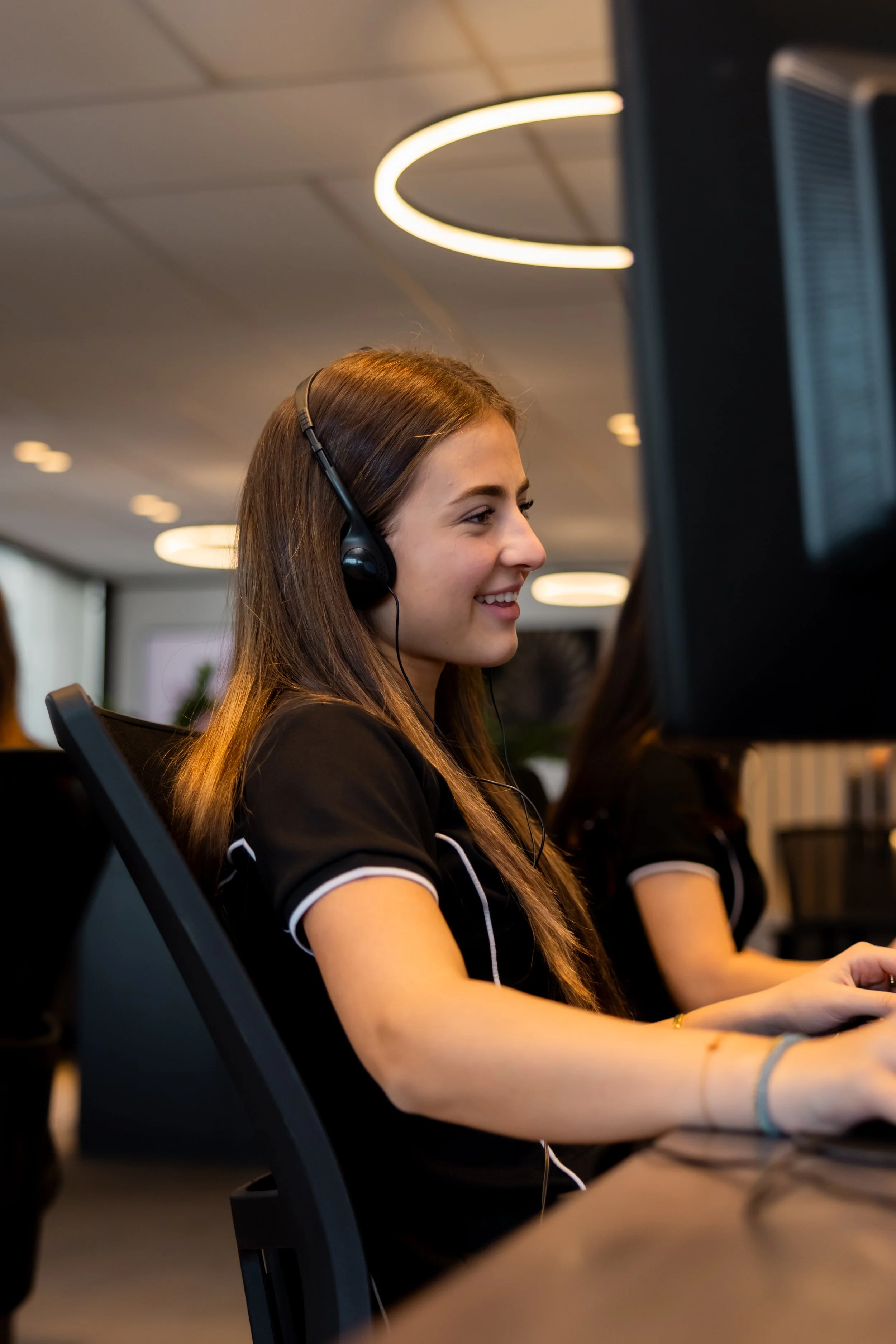 A young woman with long brown hair wearing a black shirt with white trim, headphones, and a bracelet, working on a computer in an office environment with ceiling lights.