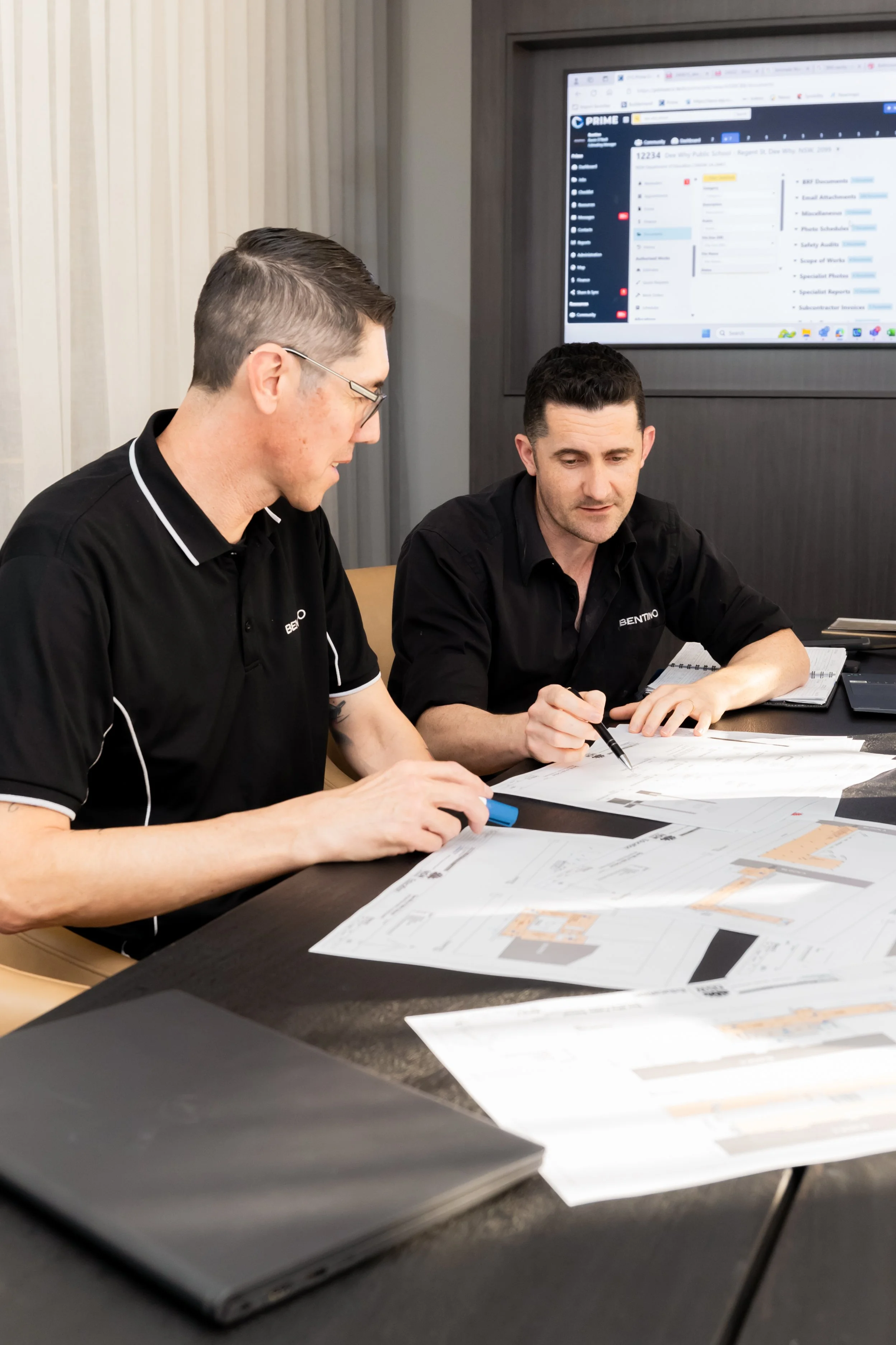 Two men in black collared shirts sitting at a table with architectural plans, reviewing documents, with a large computer monitor in the background displaying a project management software.