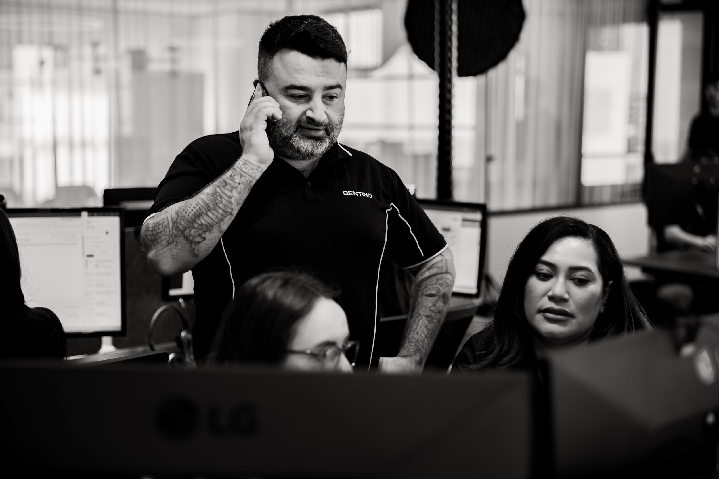 A man with tattoos on his arm and a beard, wearing a shirt with the name 'BENTINO,' is talking on the phone while standing in an office environment with multiple computer monitors and large windows in the background.